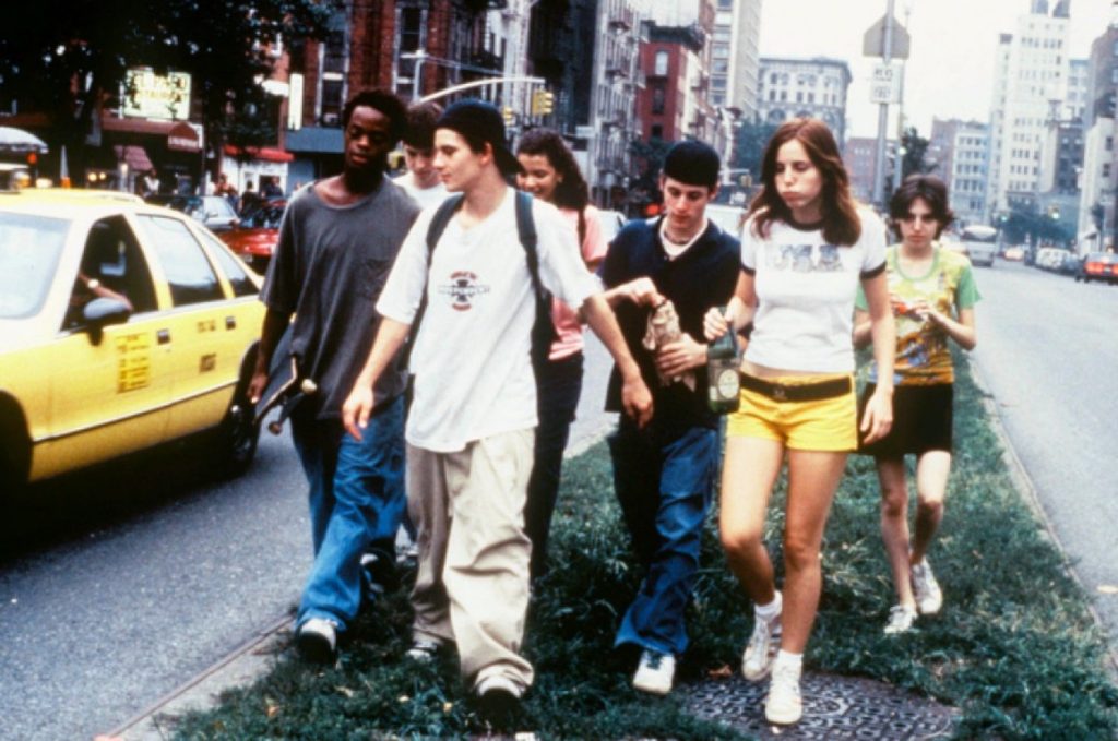 Film still from Kids showing a group of teenagers walking along a city street beside traffic, with buildings and a yellow taxi in the background.